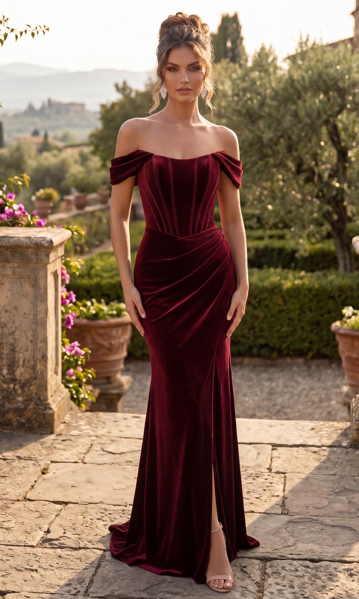 A woman stands outdoors on stone pavement, surrounded by lush greenery and blooming flowers, wearing the Chicas DM809 Long Velvet Prom Dress in burgundy with an off-shoulder design and a corset bodice.
